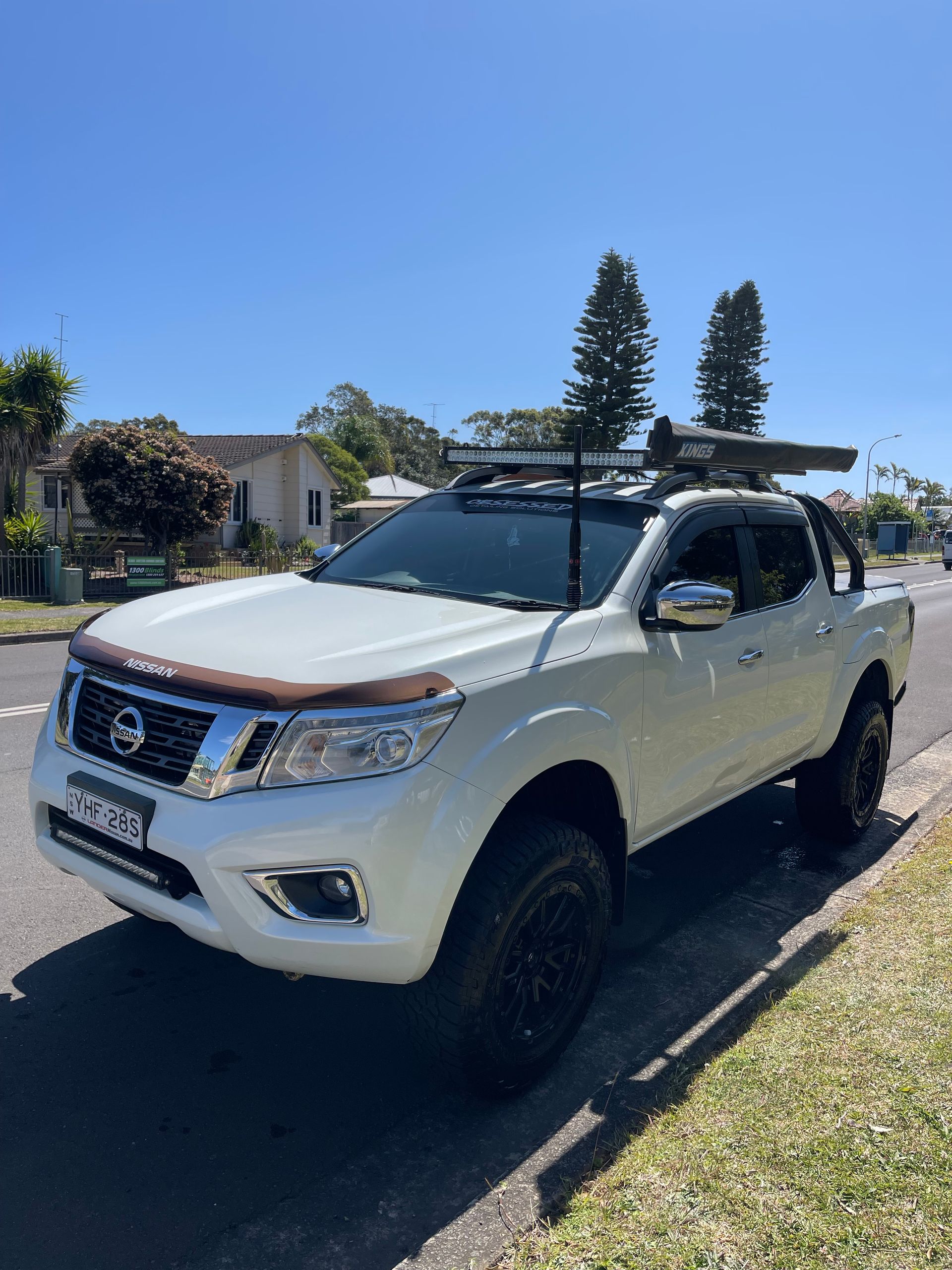 White Nissan Navara parked on a residential street with a roof rack, snorkel, and black off-road wheels.