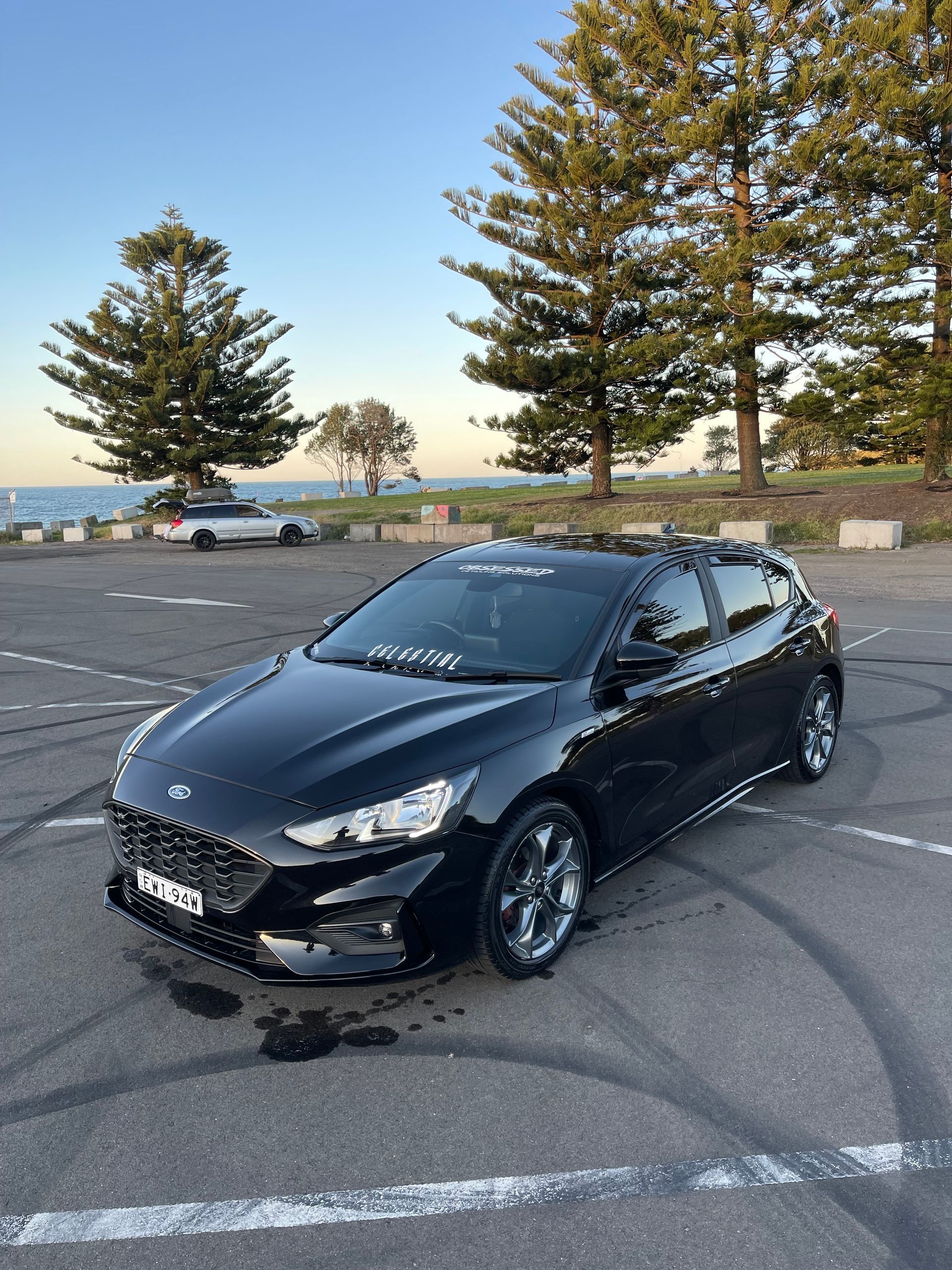 Black Ford hatchback parked on asphalt with tire tracks near a body of water and trees.