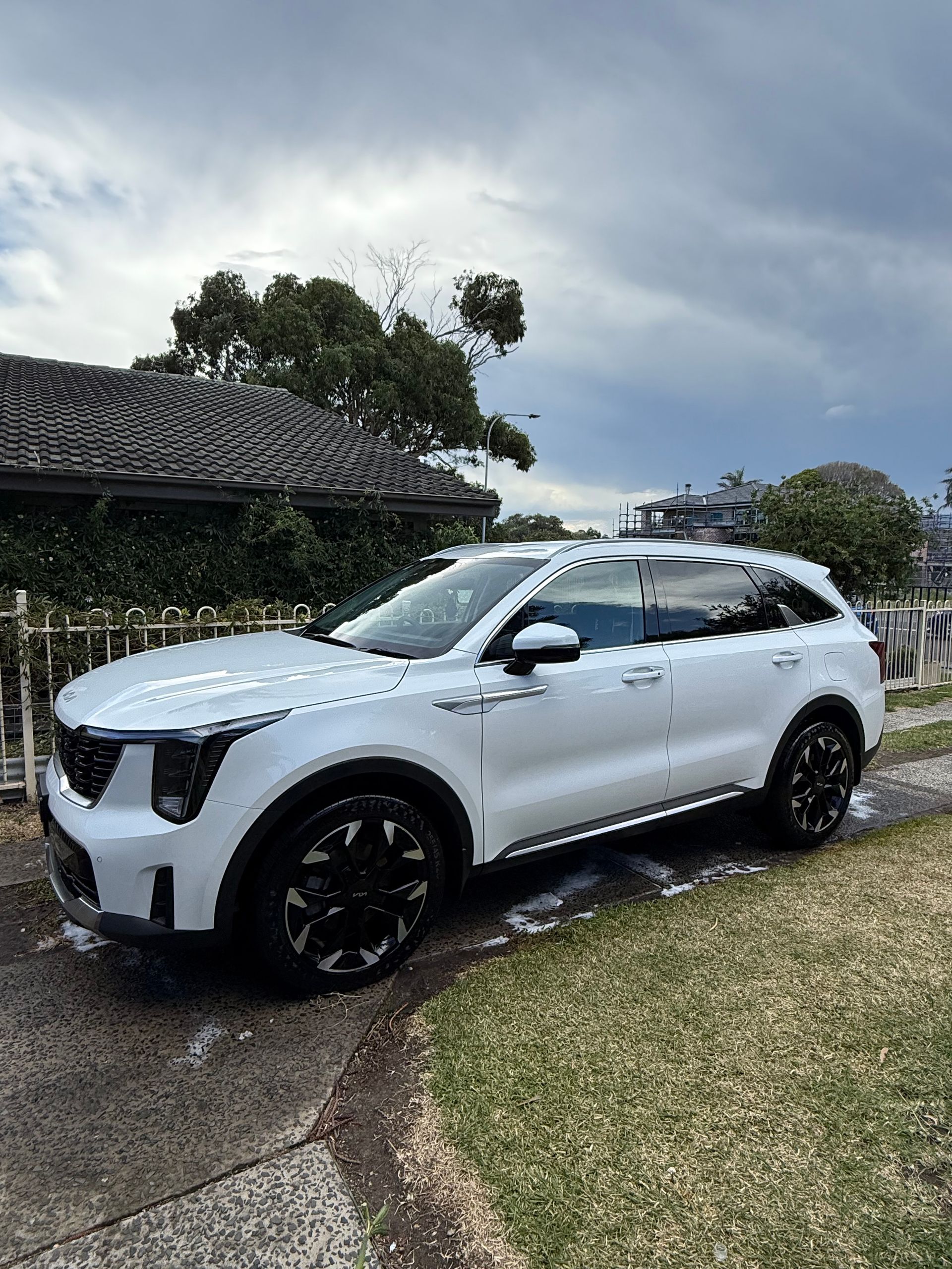 White Kia SUV parked on a driveway with black wheels, green grass, and cloudy sky background.