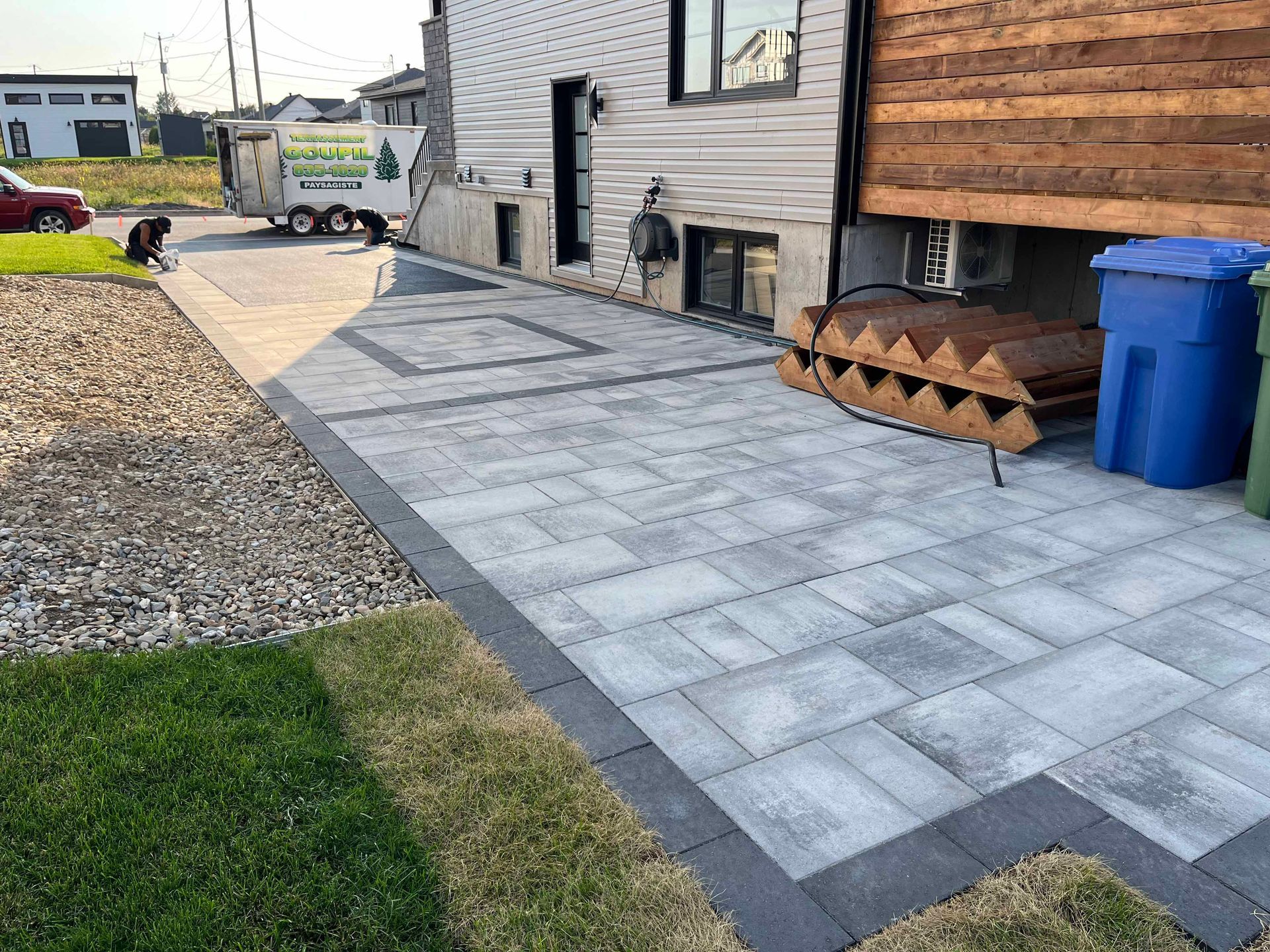 Une terrasse en pavés gris récemment installée, bordée d'une frise foncée, longe une maison d'habitation aux murs revêtus de bois et de bardage clair.