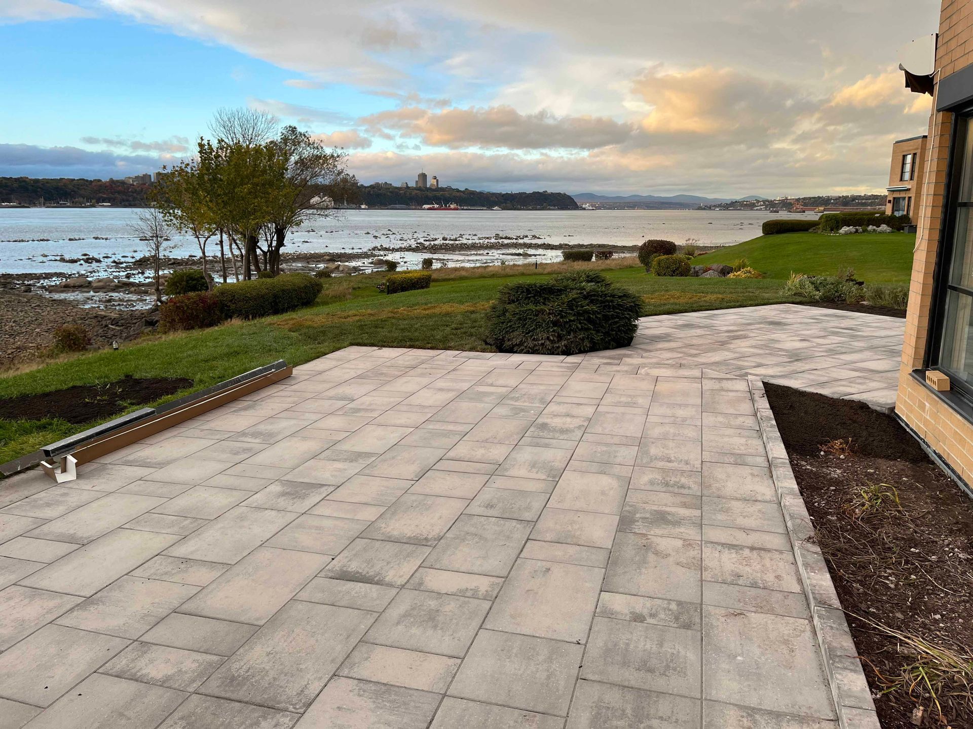 Une grande terrasse rectangulaire pavée surplombe une rivière pittoresque, avec une pelouse et des arbres sous un ciel nuageux au coucher du soleil.