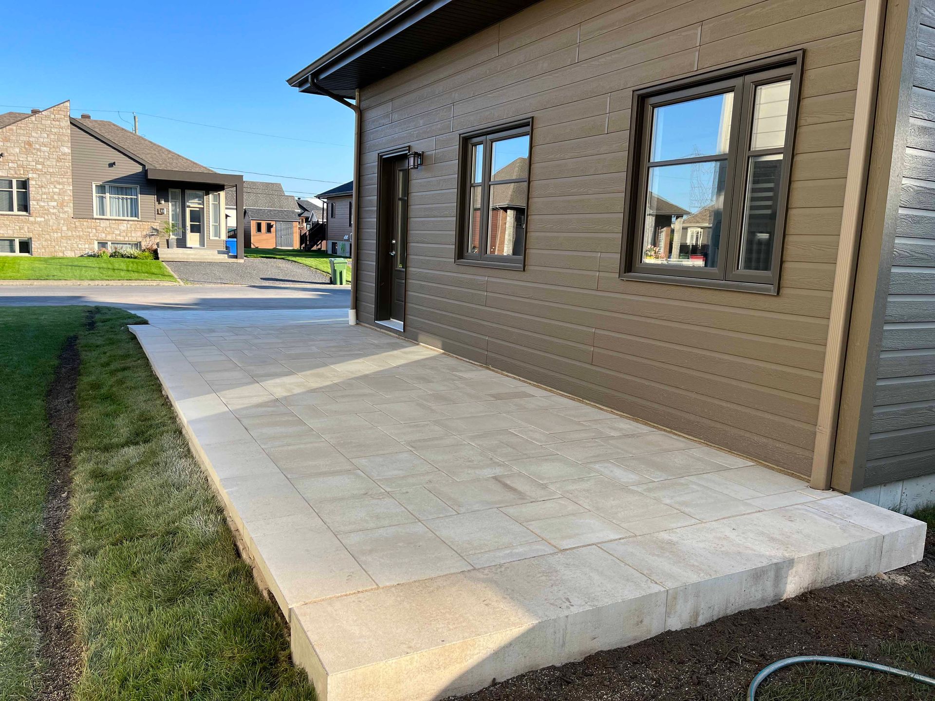 Une maison moderne au revêtement brun présente une terrasse en pierre claire jouxtant une pelouse verte sous un ciel bleu azur.