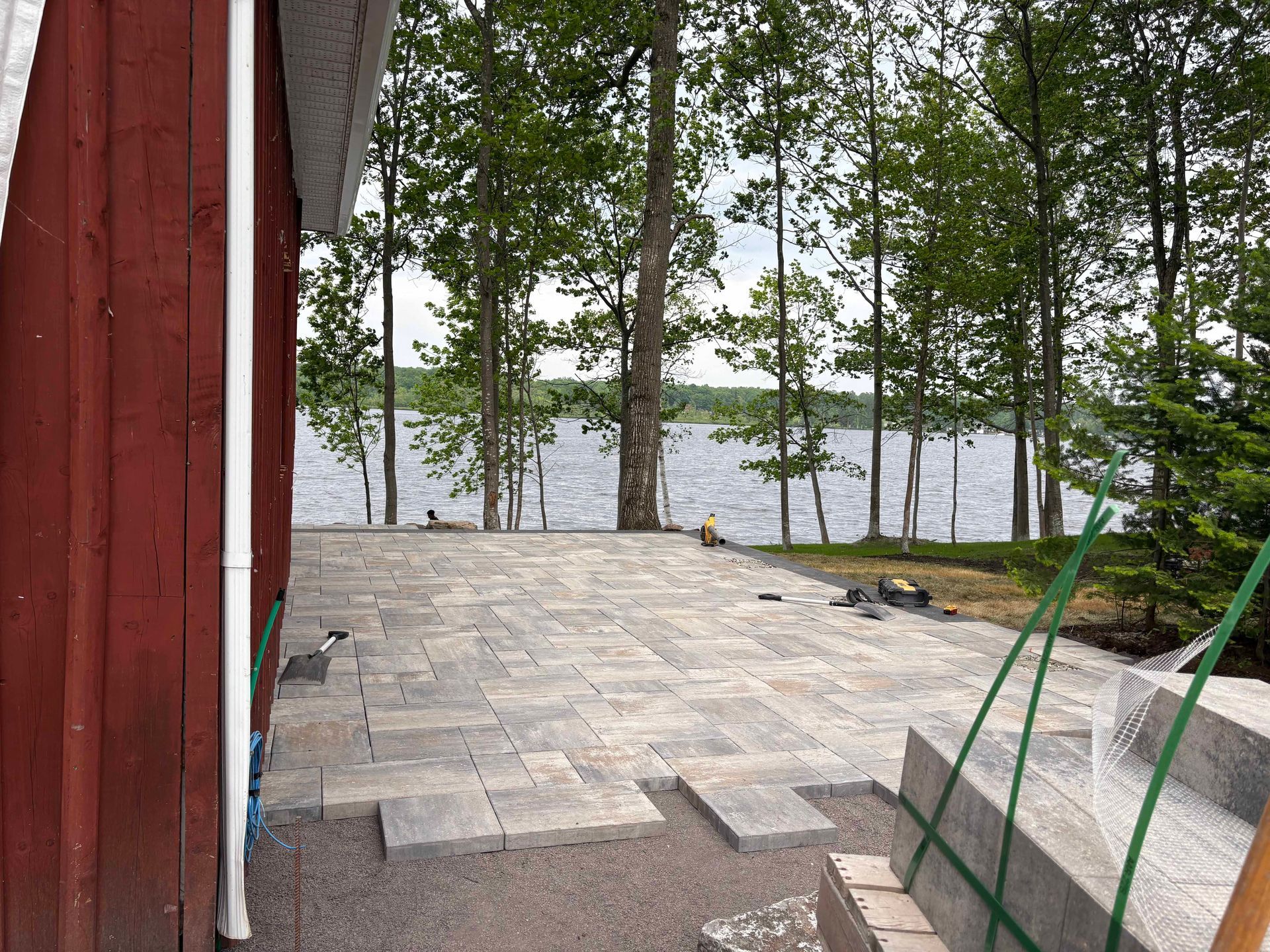 Une terrasse en pavés de pierre claire, récemment installée, jouxte un bâtiment en bois rouge, avec vue sur un lac et une forêt au-delà.