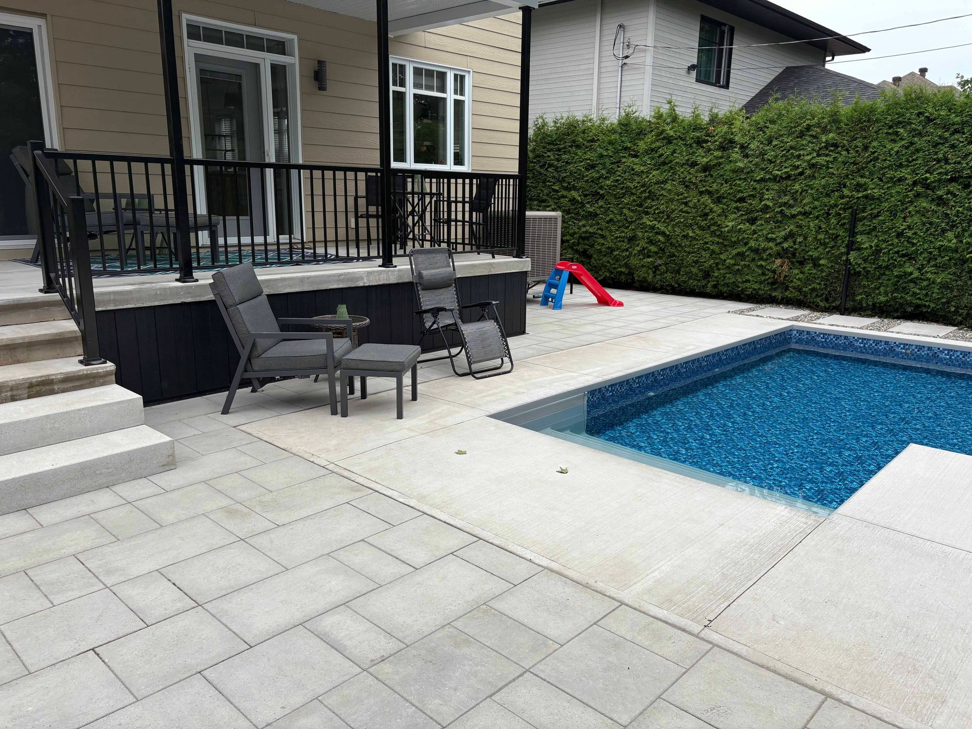 Un jardin avec des dalles de pierre gris clair, une piscine creusée, une terrasse avec des balustrades noires, deux chaises et une haie.