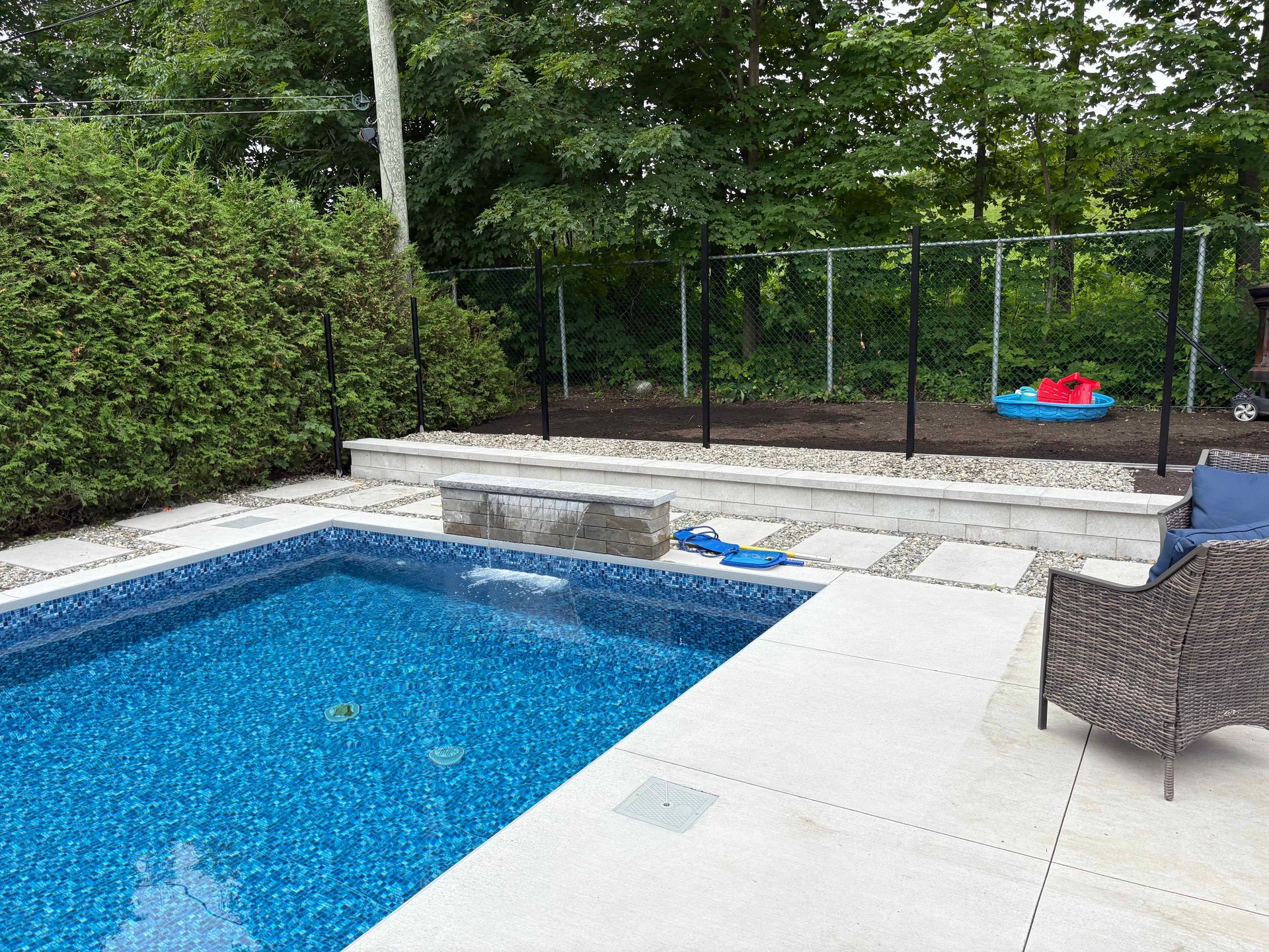 Une piscine bleue dans le jardin, agrémentée d'une fontaine en pierre, d'une terrasse pavée et d'une haie préservant l'intimité, le tout adossé à un jardin clôturé.