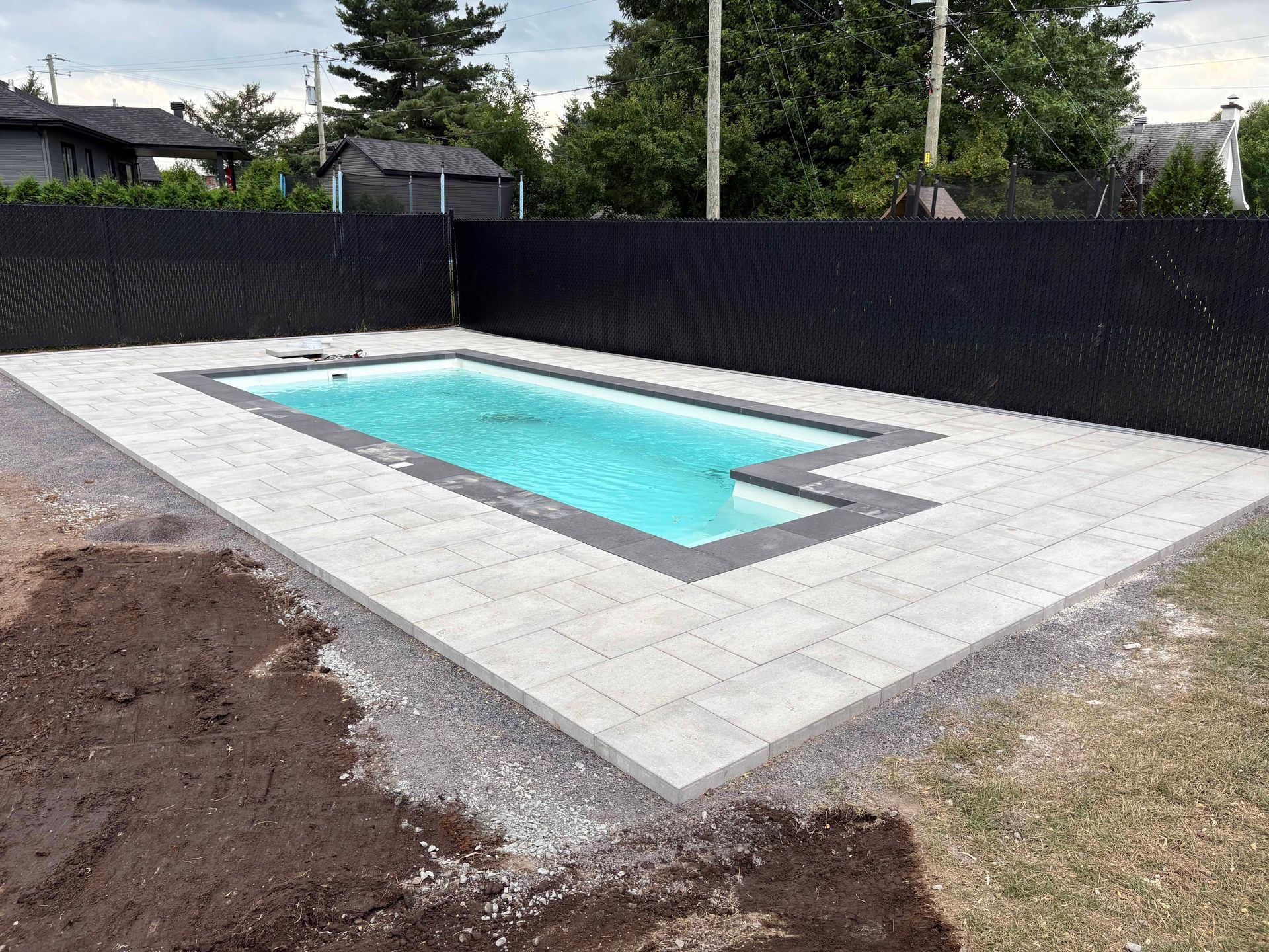 Une piscine rectangulaire à l'eau bleu clair et une terrasse en pavés gris, entourée d'une haute clôture noire assurant l'intimité.