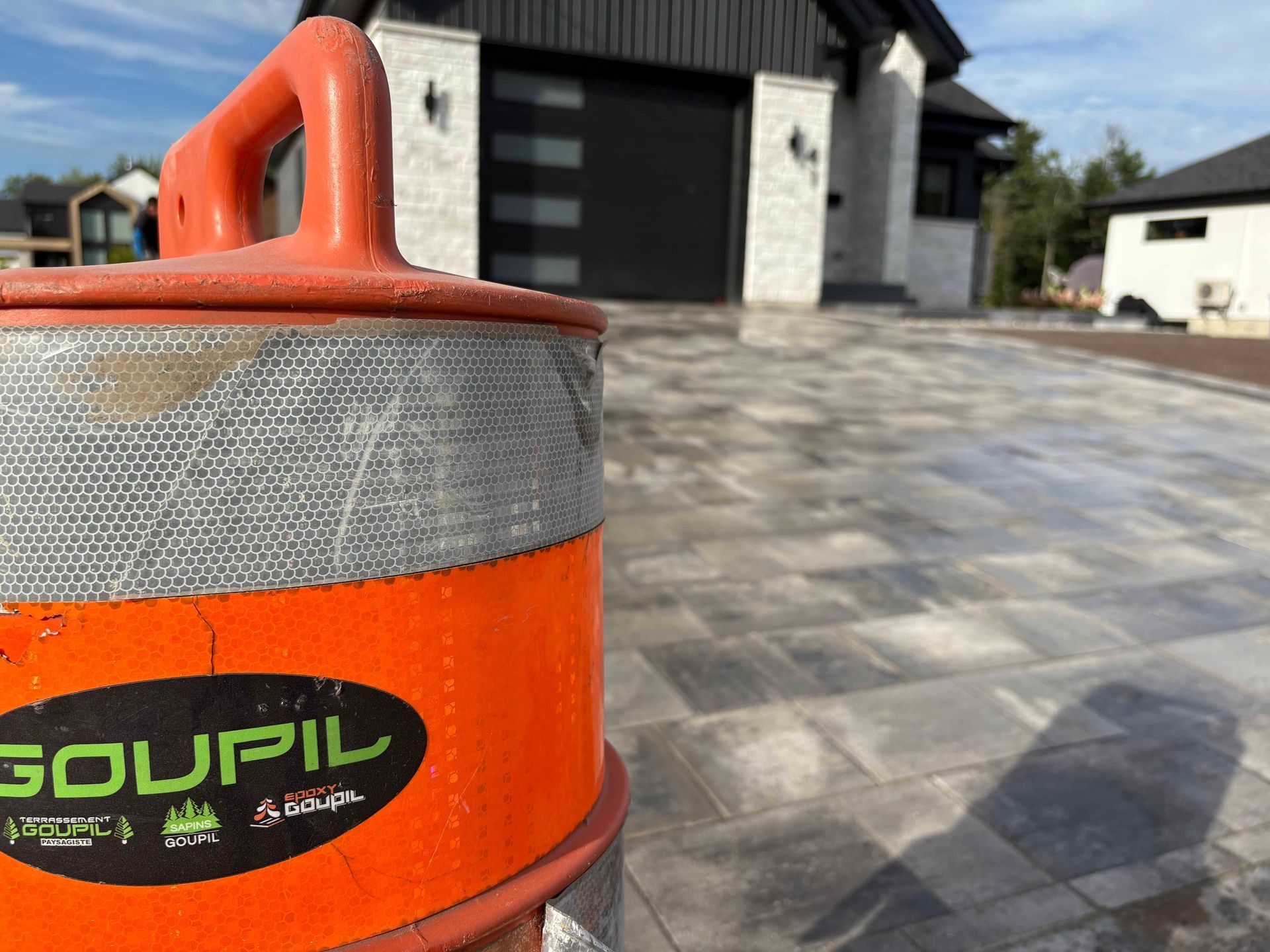 Gros plan sur un fût de construction orange Goupil au premier plan d'une maison moderne avec une allée pavée.