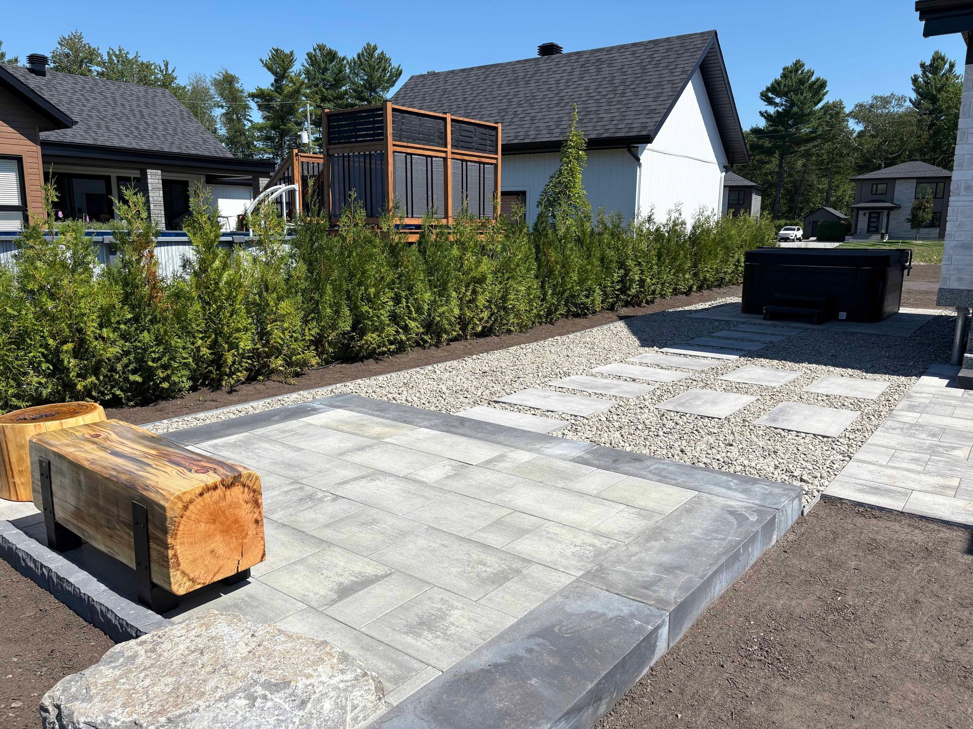 Une terrasse paysagée à l'arrière de la maison, avec un banc en rondins, des dalles de pierre claire, une zone de gravier, une haie et un jacuzzi noir.