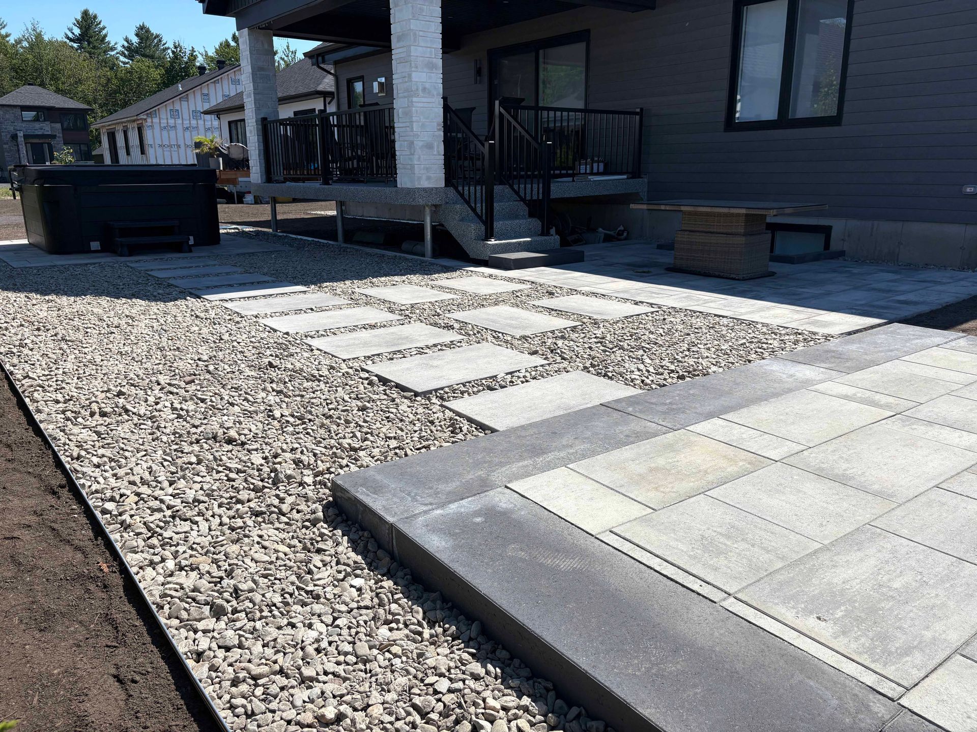 La terrasse arrière est composée de pavés carrés gris clair bordés d'une bordure en pierre plus foncée, adossée à une zone de gravier et au porche de la maison.