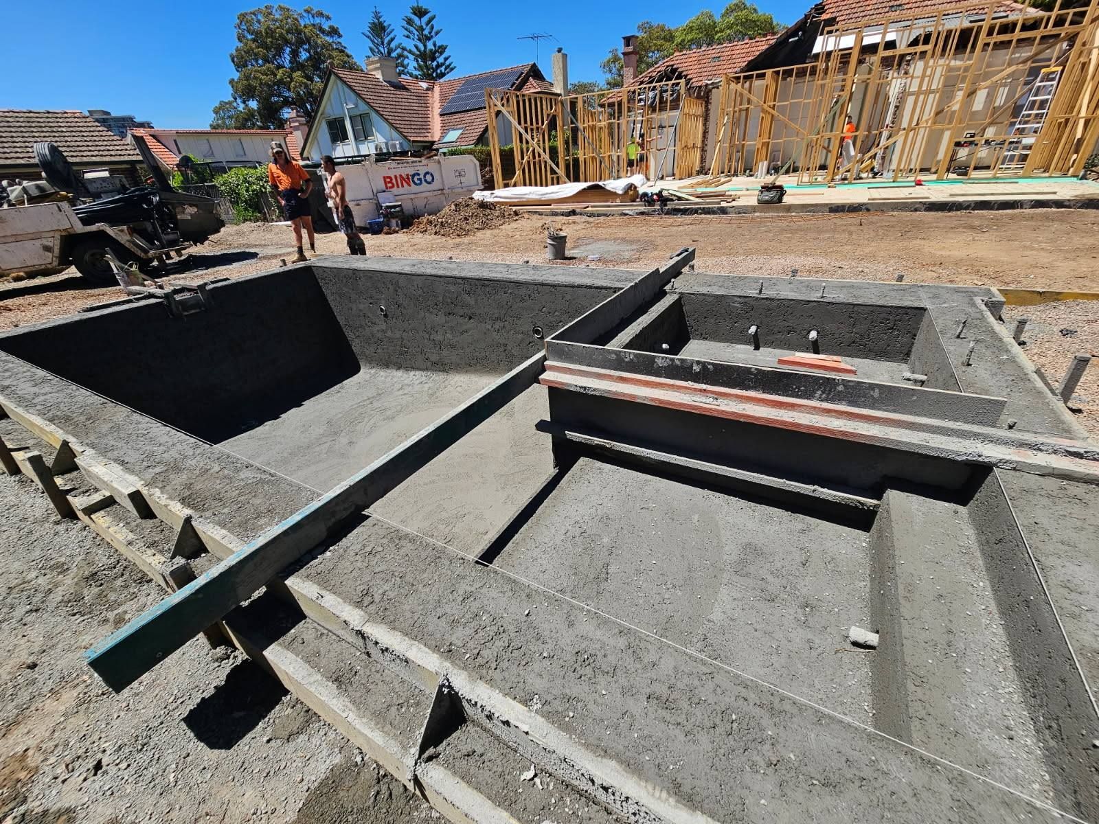 Concrete foundation under construction with wooden framing and workers on a sunny residential site