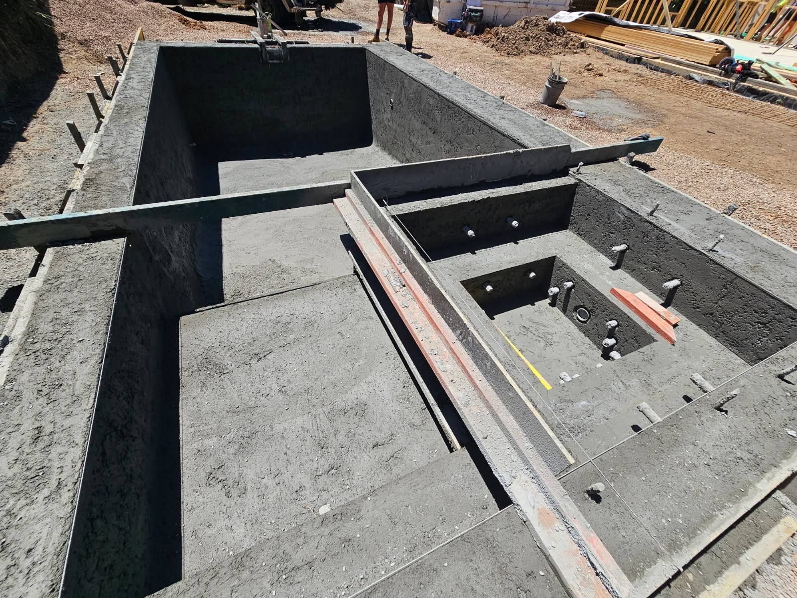 Construction site with concrete foundation trenches and a rectangular pit with metal and wood framing.