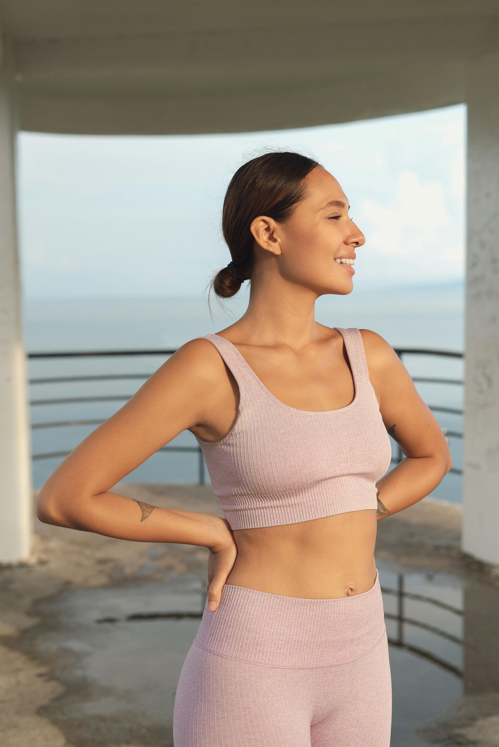 Woman in pink workout set smiling, looking to the side, hands on hips, near a body of water.