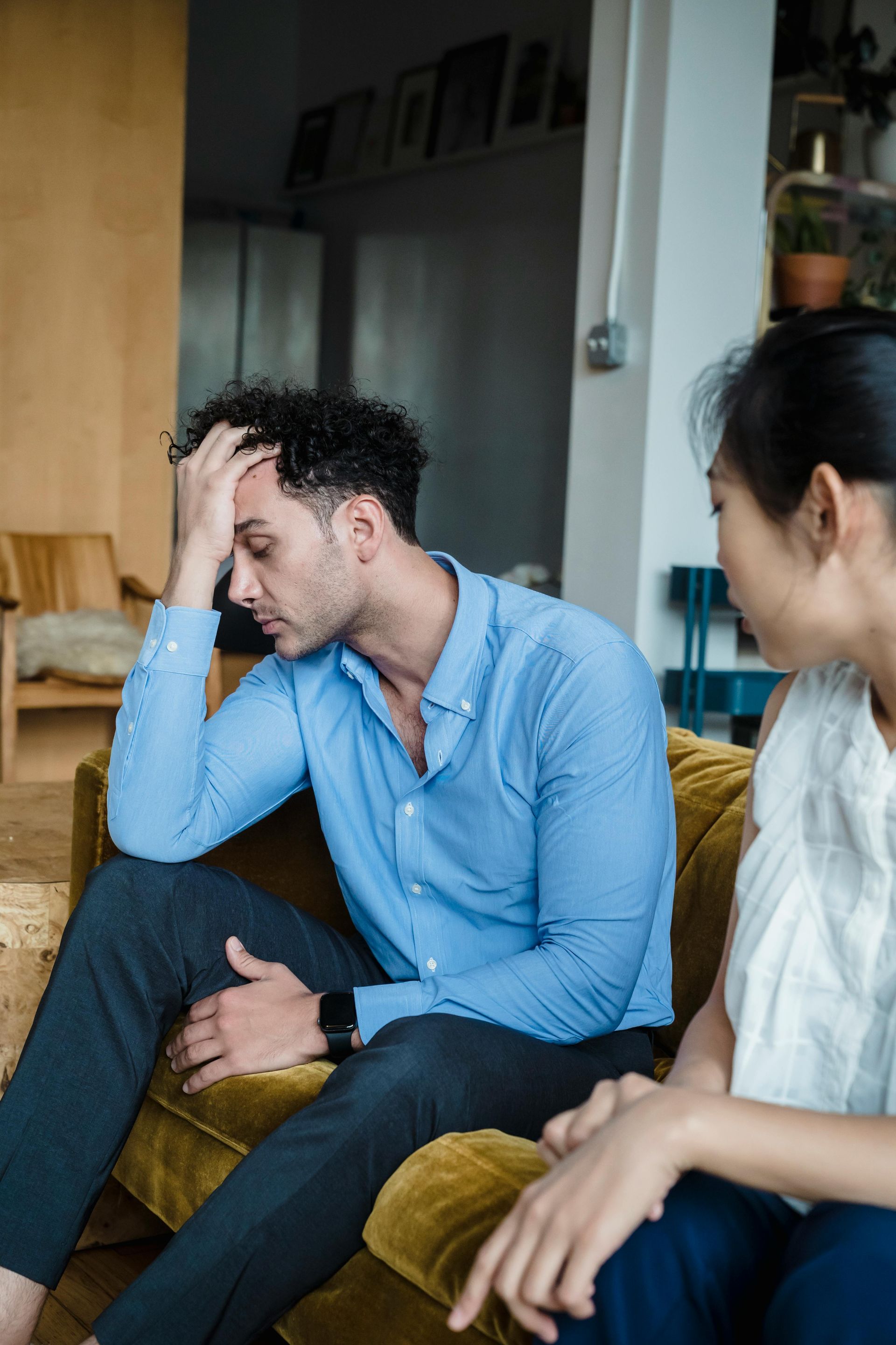 Man with head in hand, looking stressed, sits on a sofa. Woman beside him appears concerned.
