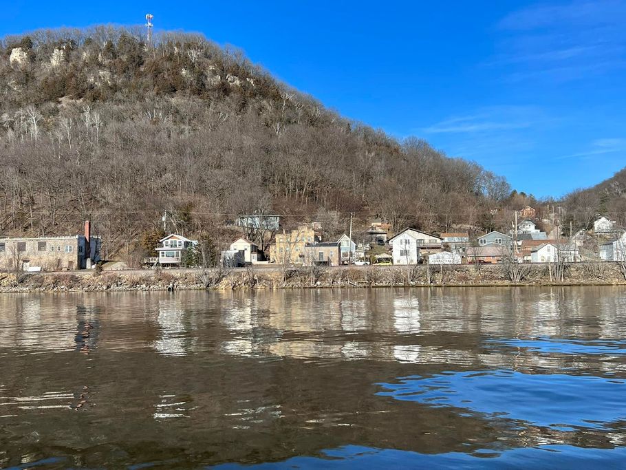 Town nestled at base of a wooded hill, reflected in a wide river under a blue sky.