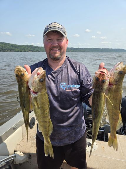 Man on a boat holding up four large fish he caught, with water and shoreline in background.