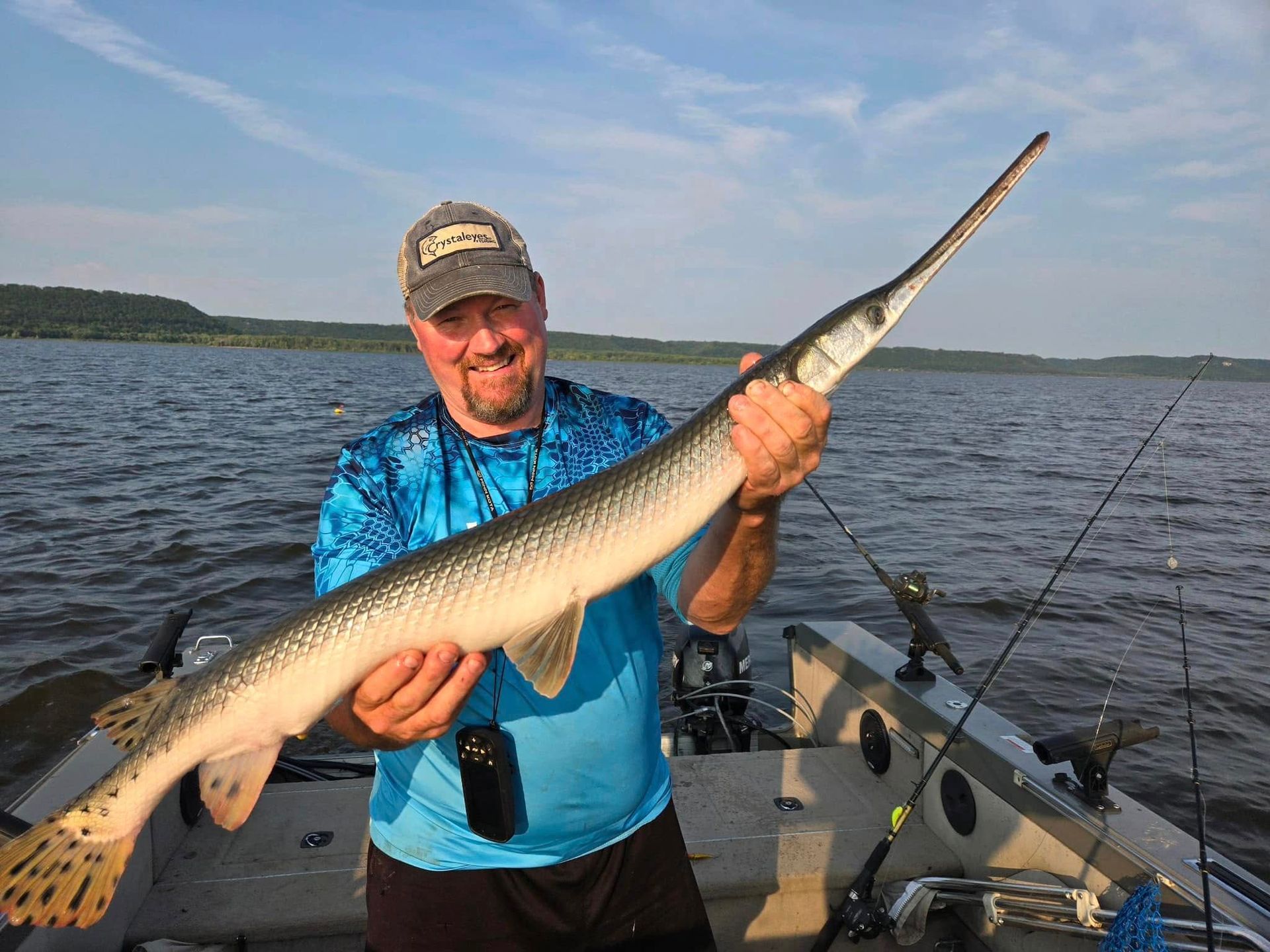 Man smiles, holding a longnose gar fish on a boat, against a lake backdrop.