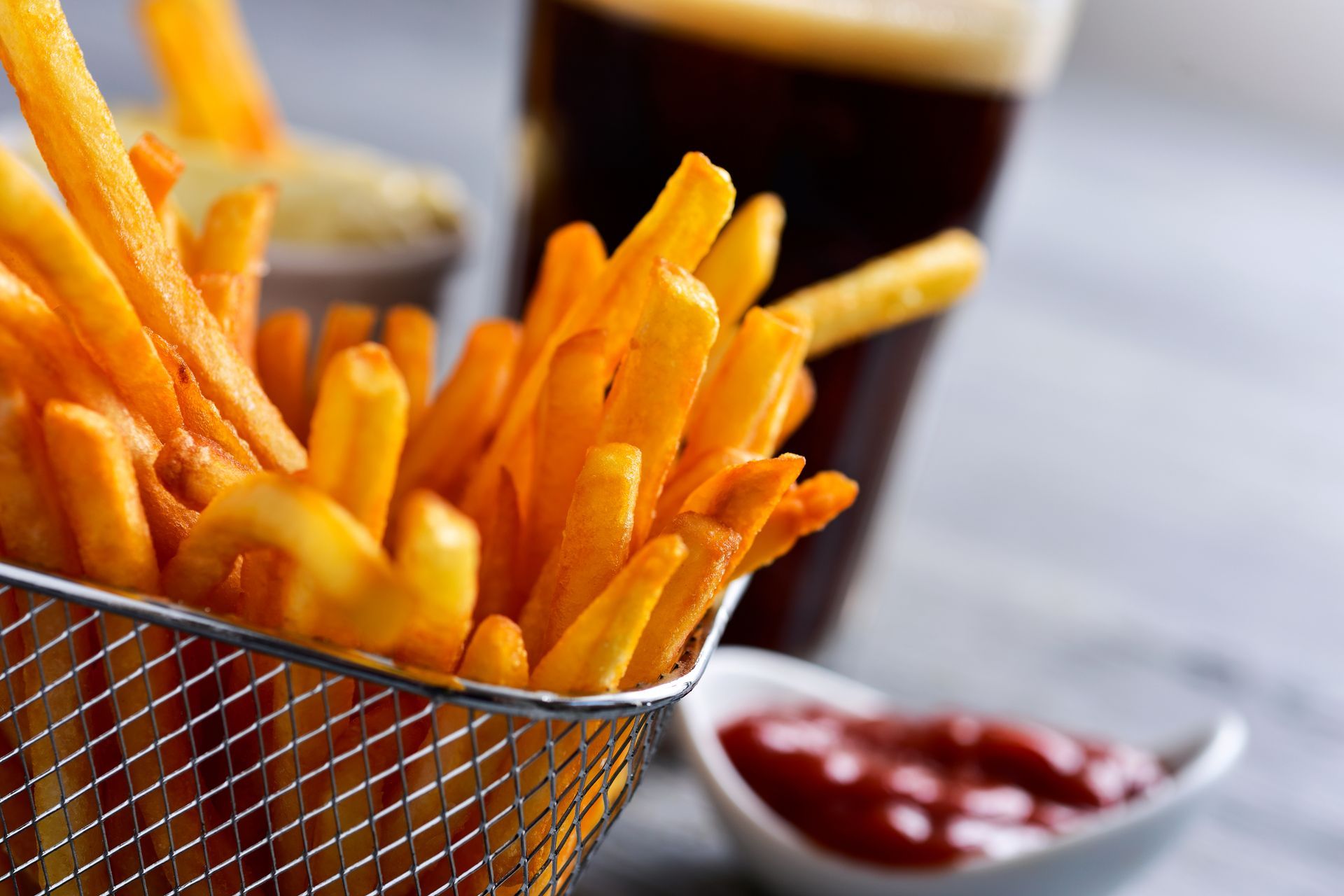French fries in a metal basket, with ketchup and a dark beverage in the background.
