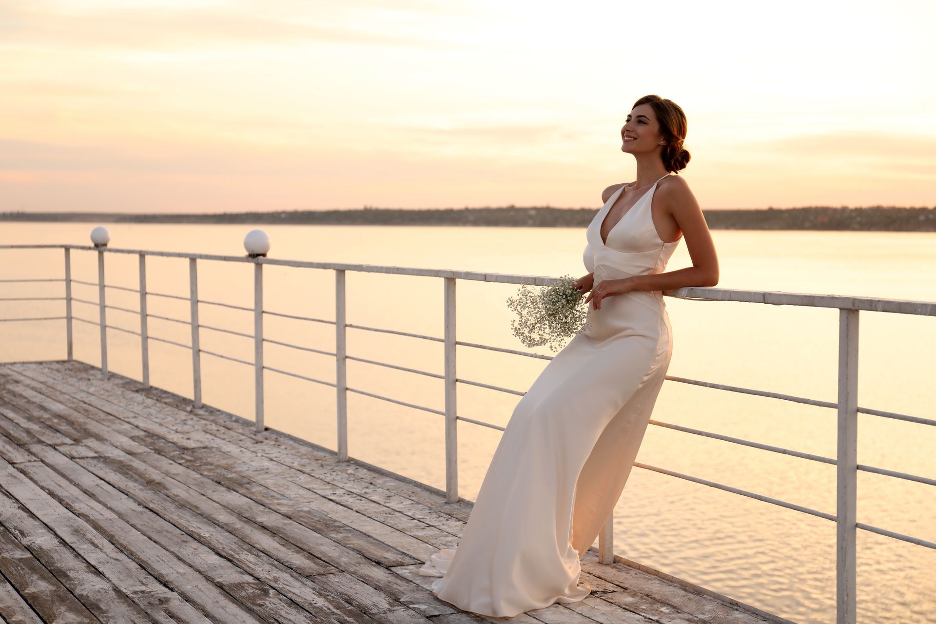 Woman in white dress leans on pier railing, holding flowers, gazing at sunset over water.