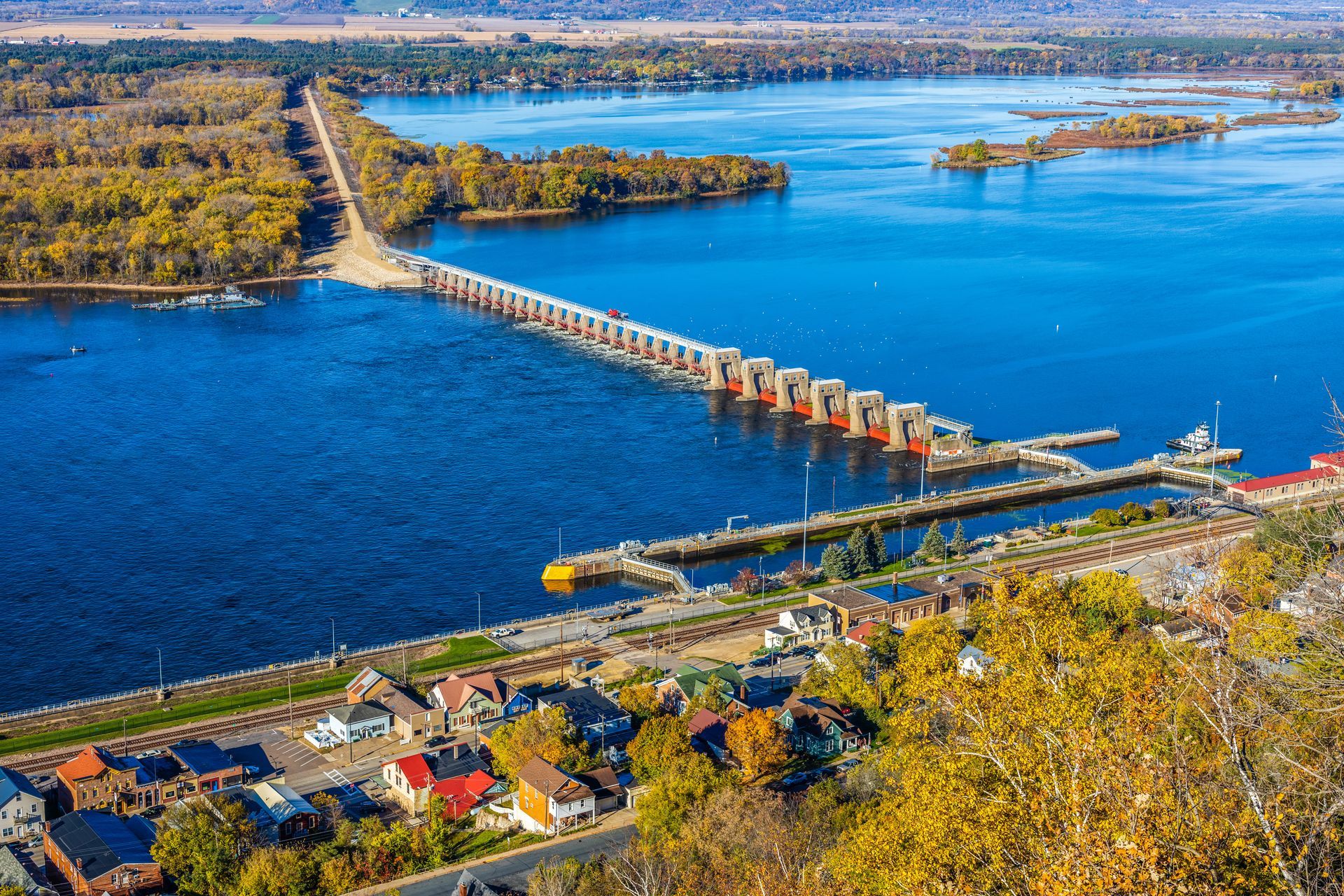 Aerial view of the Mississippi River with Lock and Dam 3, autumn trees, and colorful houses along the riverbank.