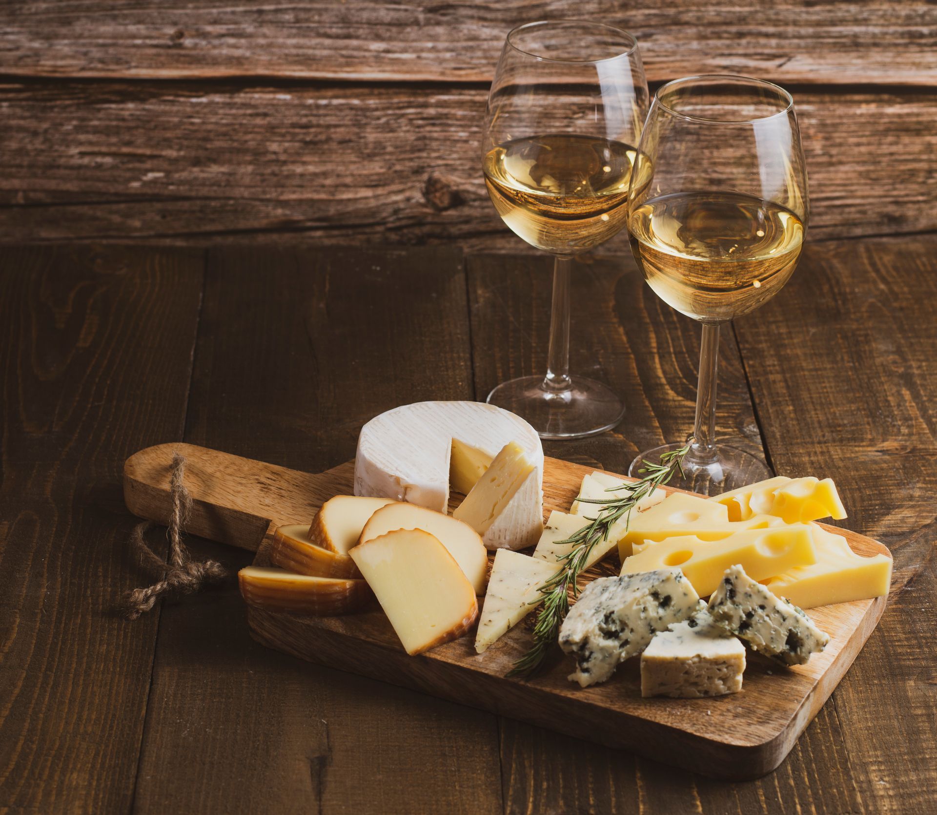 Cheese and crackers arranged on a wooden board with two glasses of white wine on a rustic wooden table.