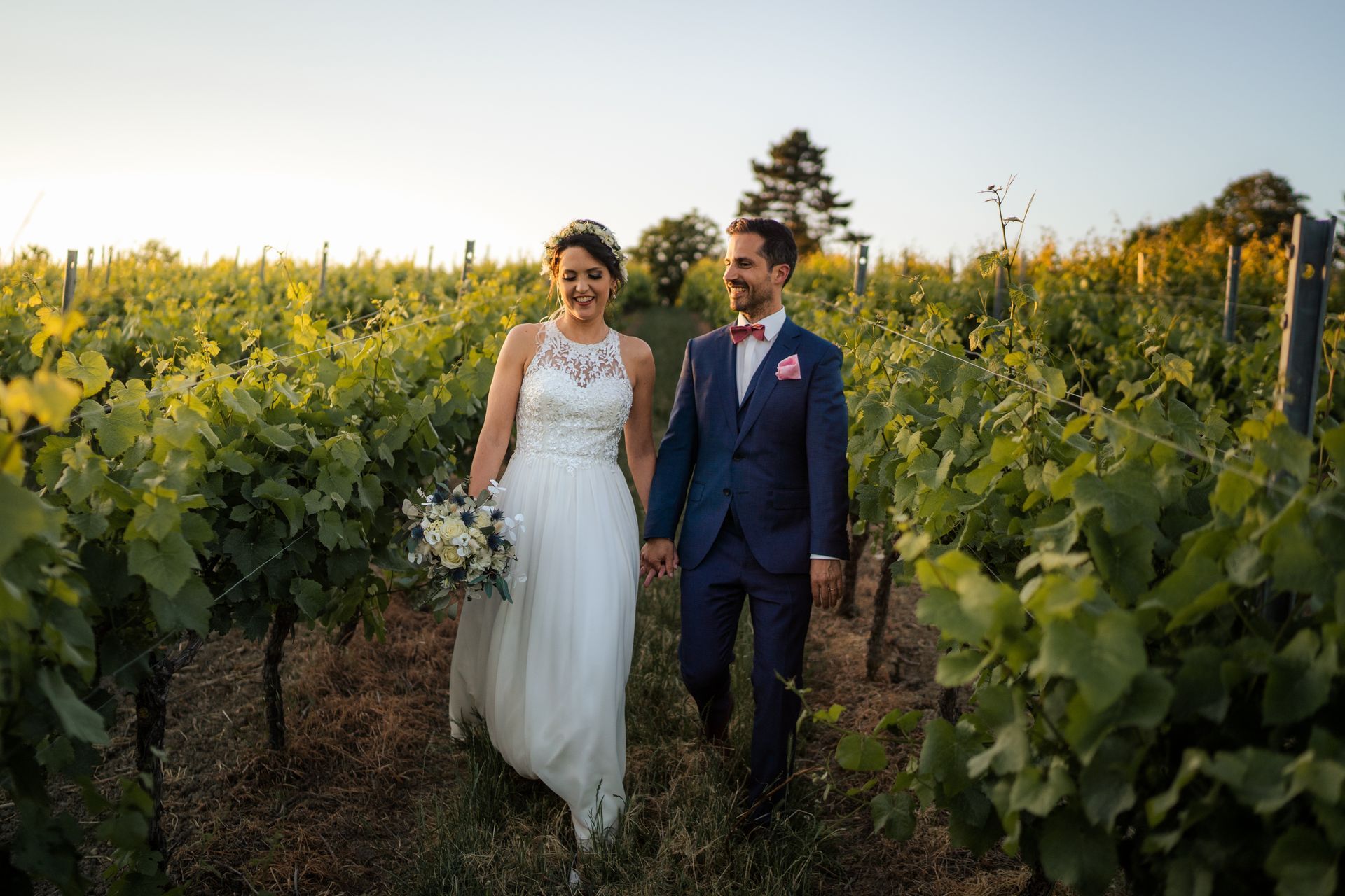 Newlyweds walk hand-in-hand through a vineyard. The woman wears a white gown; the man, a blue suit.