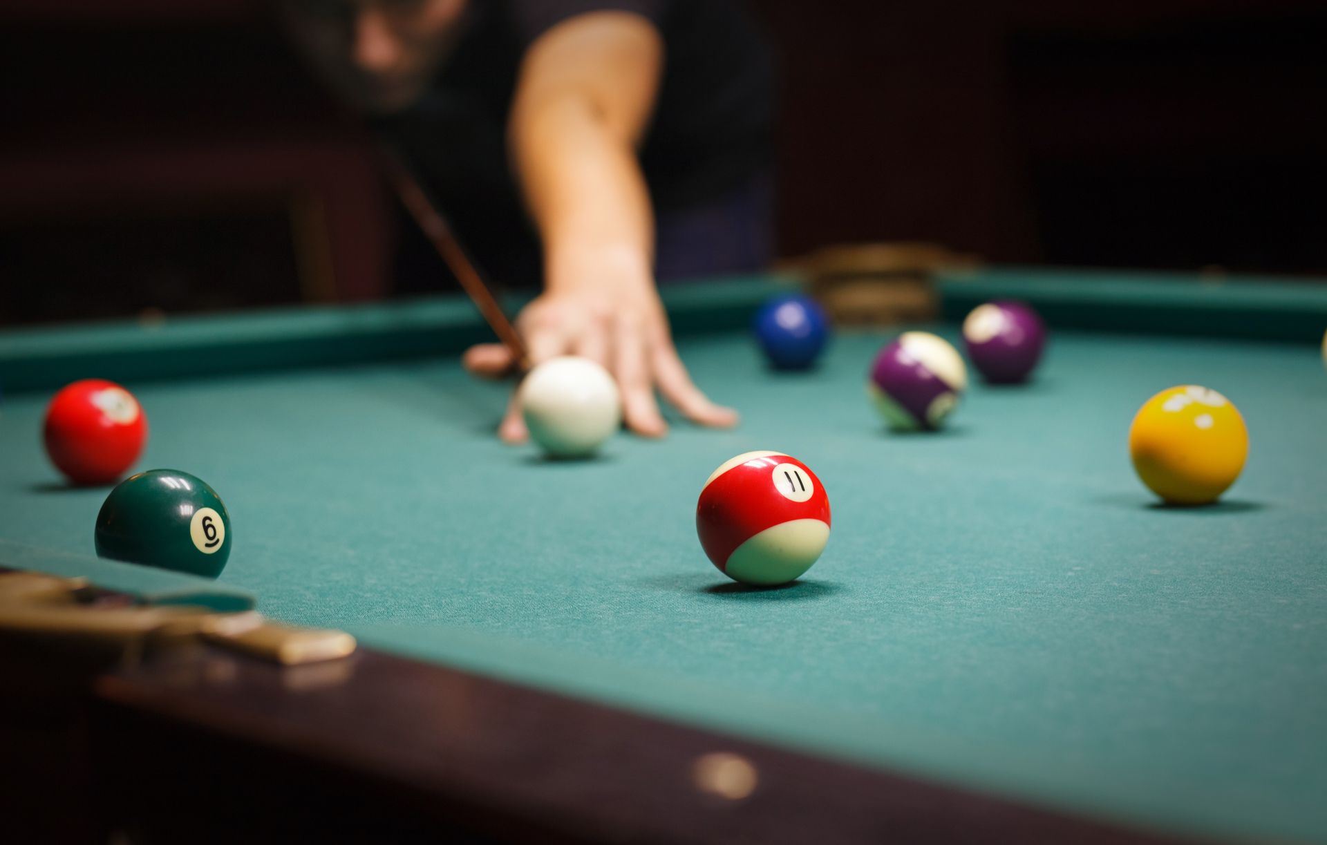 Man playing pool, aiming with a cue. Colorful balls on green felt table. Low light setting.