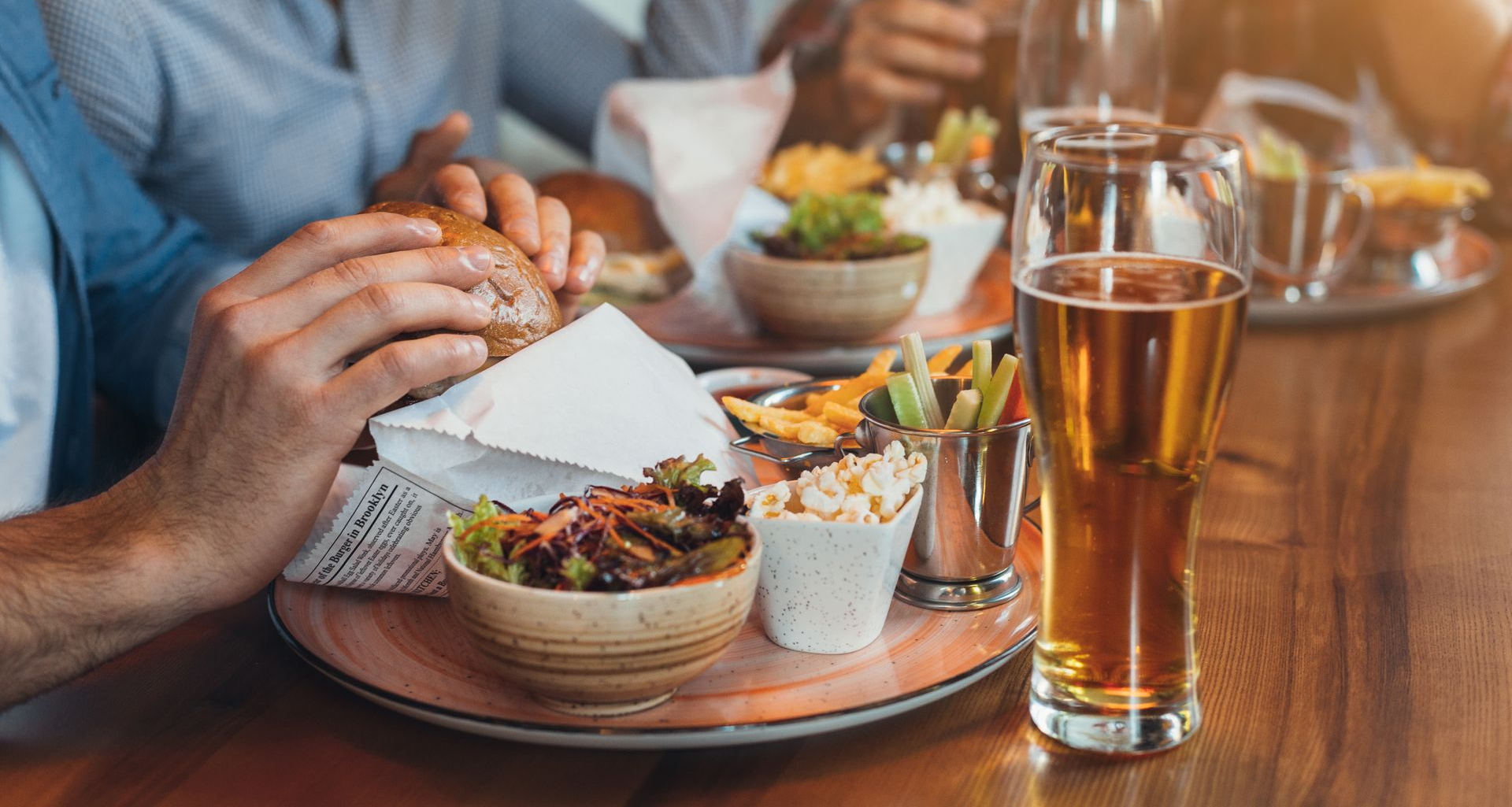 Person eating a burger with sides and beer at a wooden table in a restaurant.