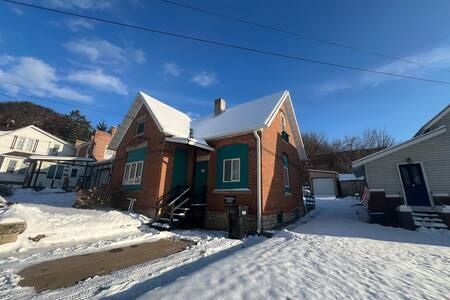 A small, one-story brick house with green trim and snow-covered roofs sits on a snowy lot under a clear blue sky.