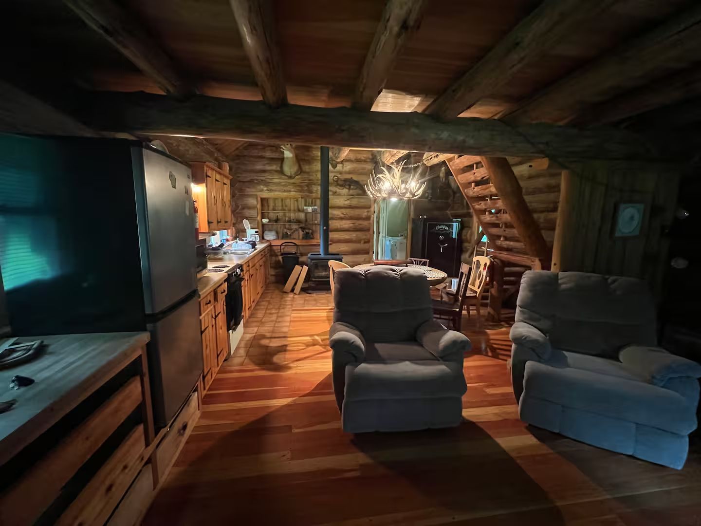 Cozy cabin interior: two recliner chairs face a dining area and kitchen with wood beams and a refrigerator.