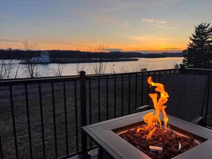 Sunset over a river, view from a deck with a lit fire pit. A barge is in the distance.