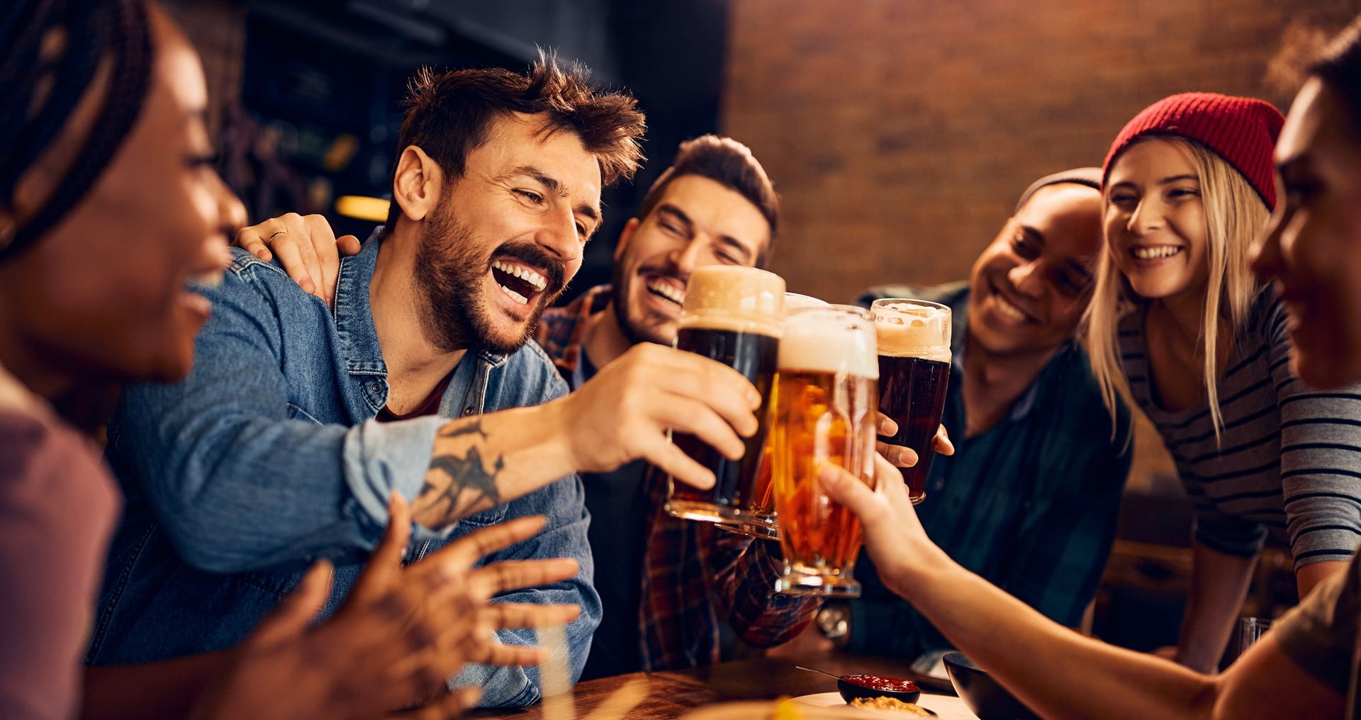 Friends at a bar, toasting beer glasses, smiling and laughing. Dark setting.