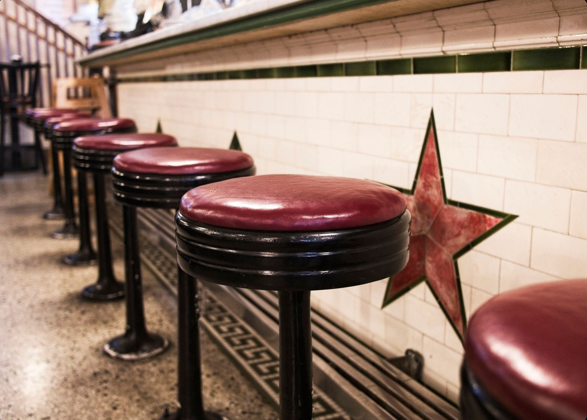 A row of red-topped bar stools along a counter with white tiled walls featuring red star patterns.