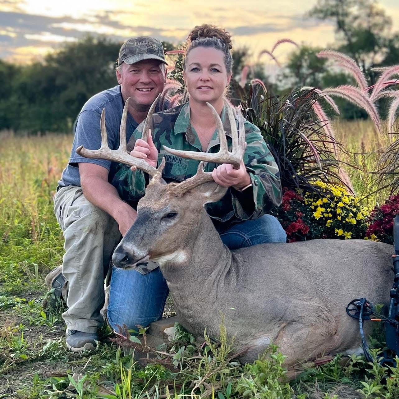 A man and woman pose with a large buck in a field. They are kneeling, smiling, and holding the deer. Autumn foliage.