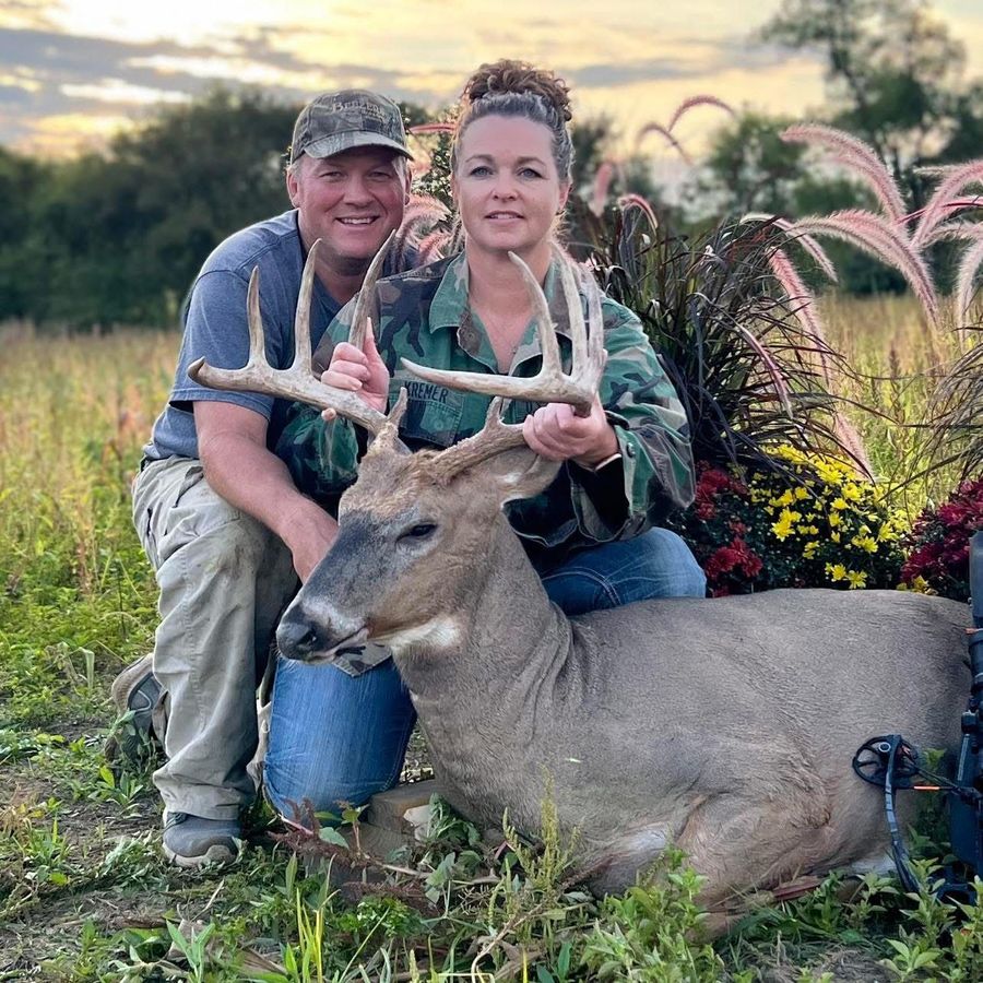 Two people kneeling with a large buck. Outdoors, field setting. Man and woman smiling, holding the buck.