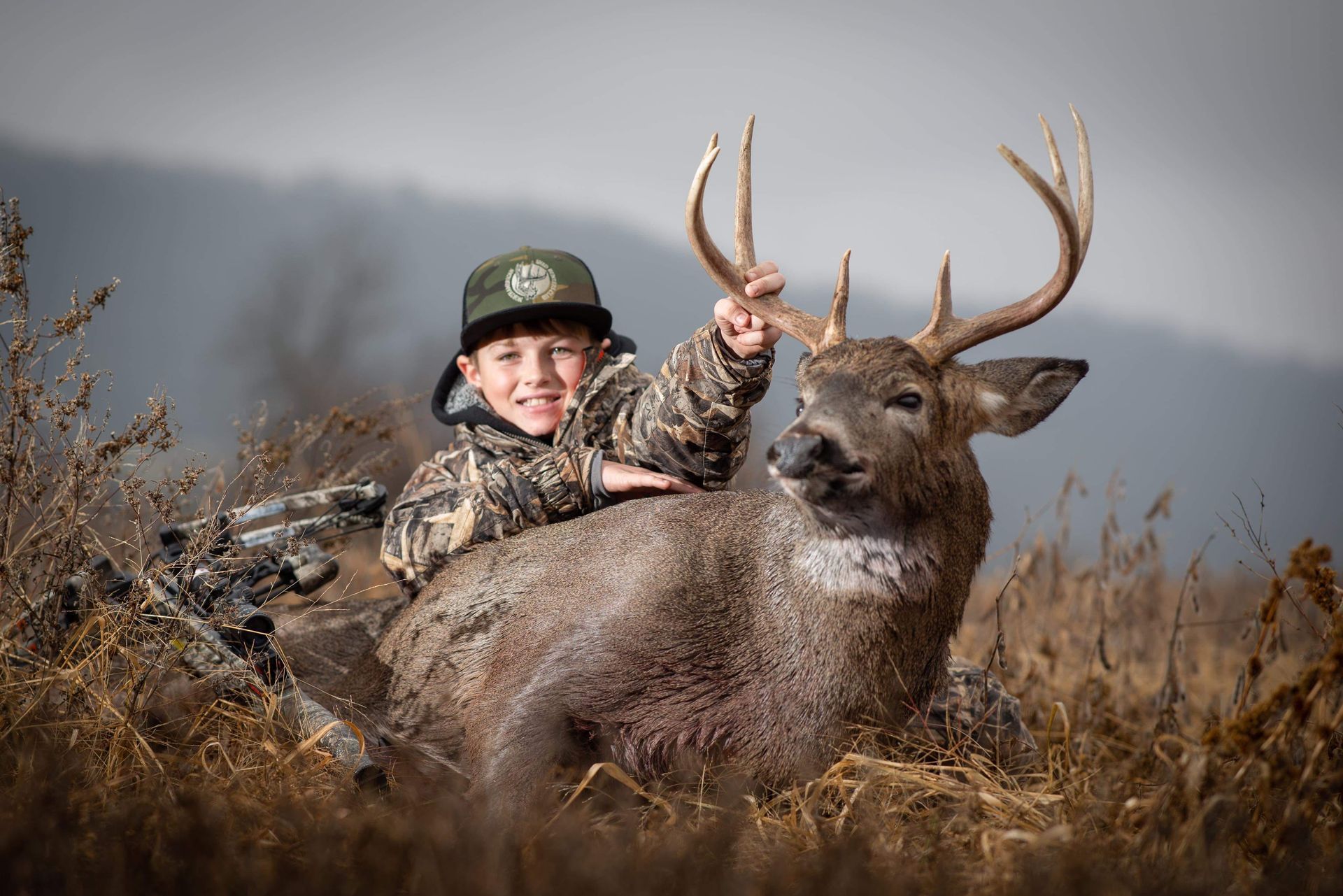 Boy in camo with a buck he harvested, outdoors.