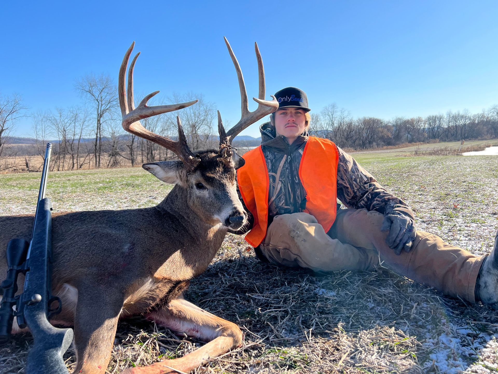 Hunter with deer, field setting. Hunter wears orange vest, poses next to large buck with rifle. Blue sky.