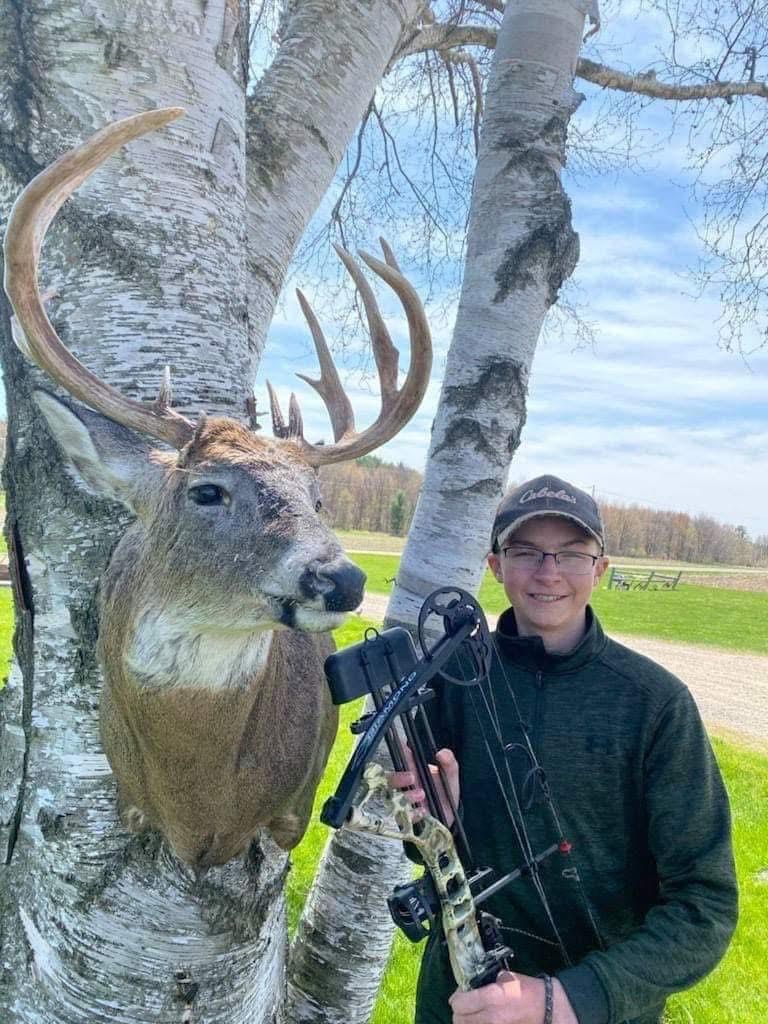 Young person with glasses, bow, and deer mount attached to birch tree.