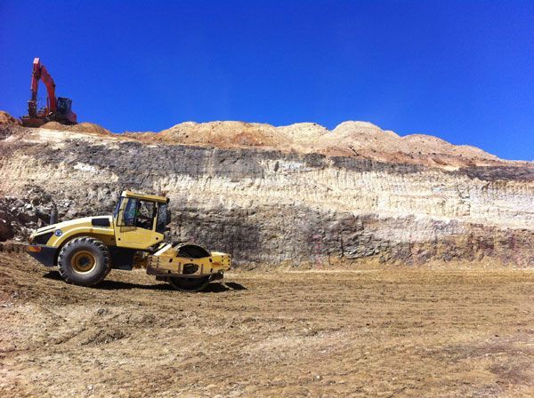 A yellow tractor is driving through a dirt field with a large excavator in the background.