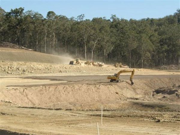 A large yellow excavator is working on a dirt field surrounded by trees.