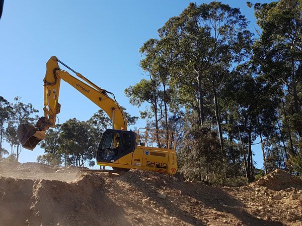 A yellow excavator is working on a dirt hill with trees in the background