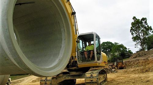 A man is sitting in a yellow excavator next to a large concrete pipe.
