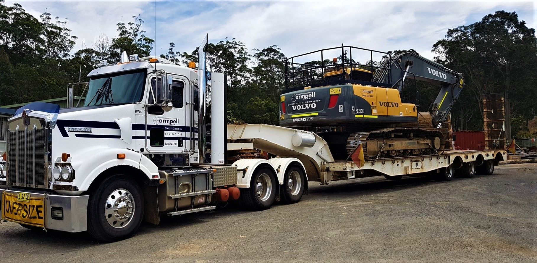 A white semi truck is carrying a yellow excavator on a trailer.