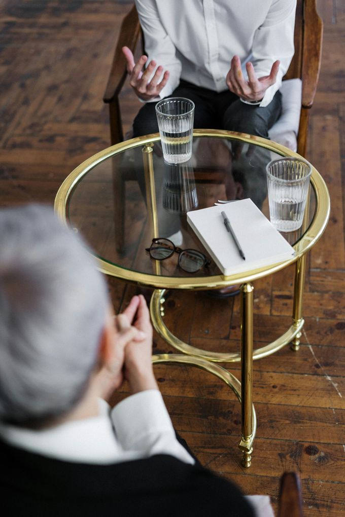 Twee mensen zitten tegenover elkaar op stoelen aan een kleine, goudkleurige glazen tafel met glazen water en een notitieblok.