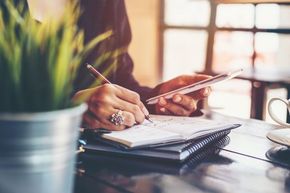 A woman is writing in a notebook while using a tablet.