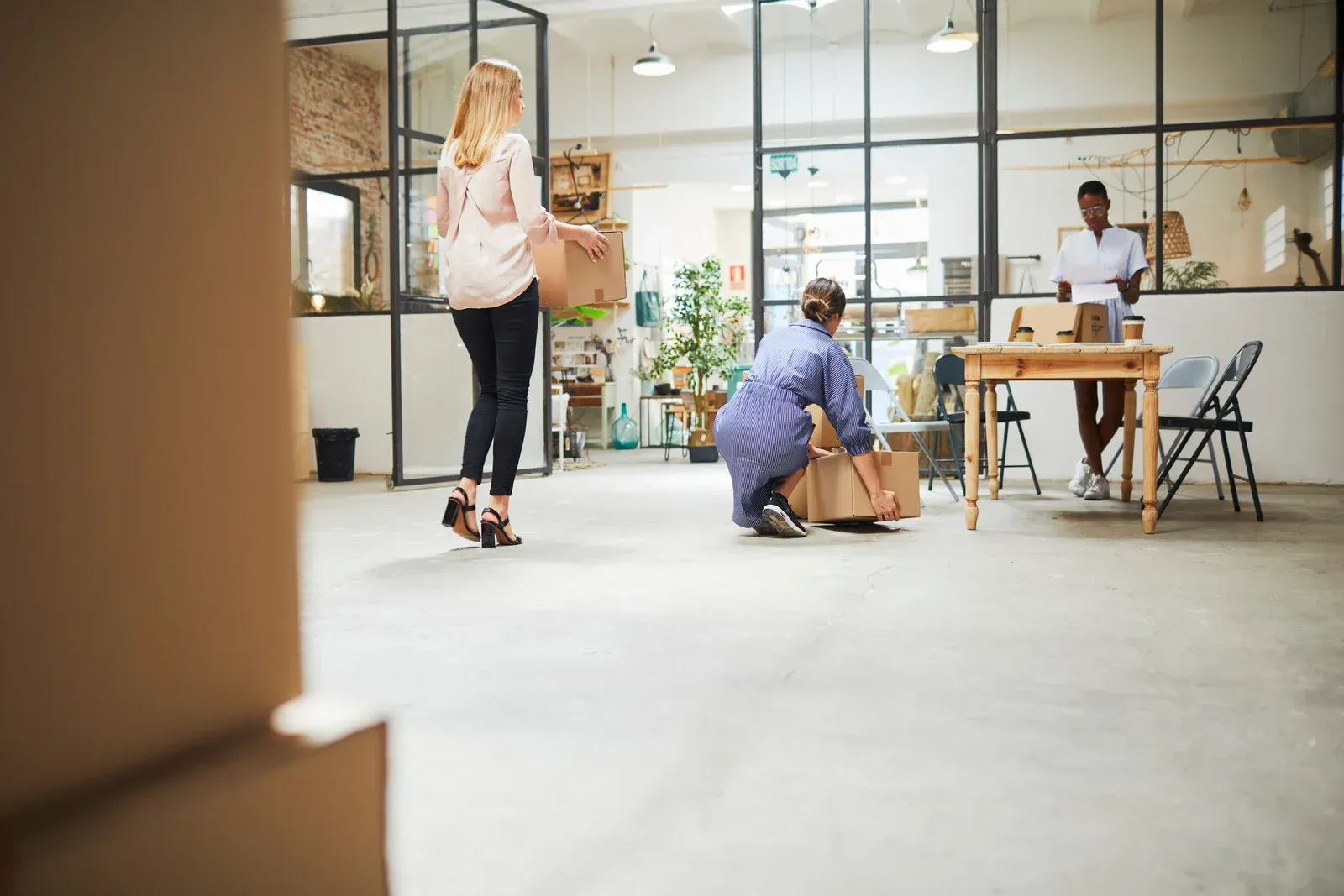 People moving boxes in a bright, modern office space. One woman carries a box; two others pack.
