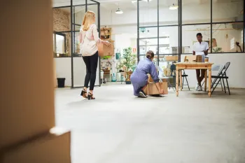 People moving boxes in a bright, modern office space. One woman carries a box; two others pack.