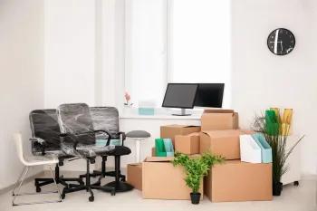 Office space being packed up; cardboard boxes stacked, chairs covered in plastic, computer monitors on desk.
