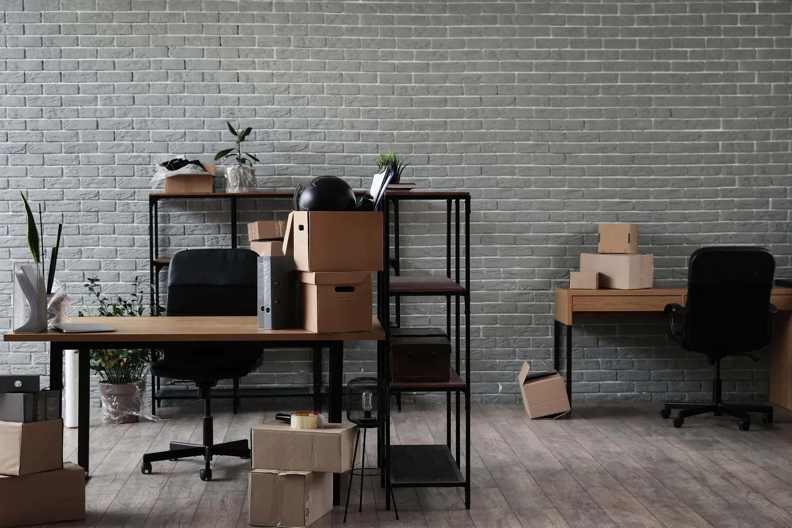 Office interior with cardboard boxes, desks, shelves, and a gray brick wall.