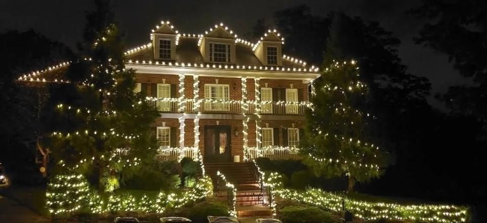 House outlined in warm white holiday lights at night, with lit trees and a glowing front porch