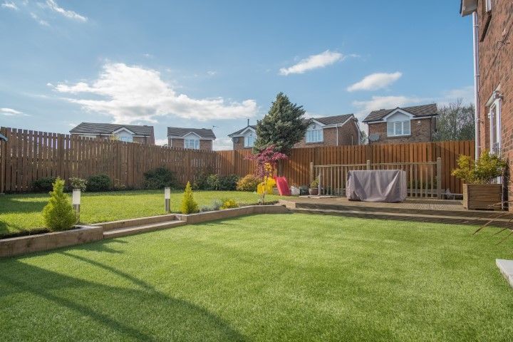 A residential backyard with green artificial grass, a wooden deck, a wooden fence, and clear blue skies above.